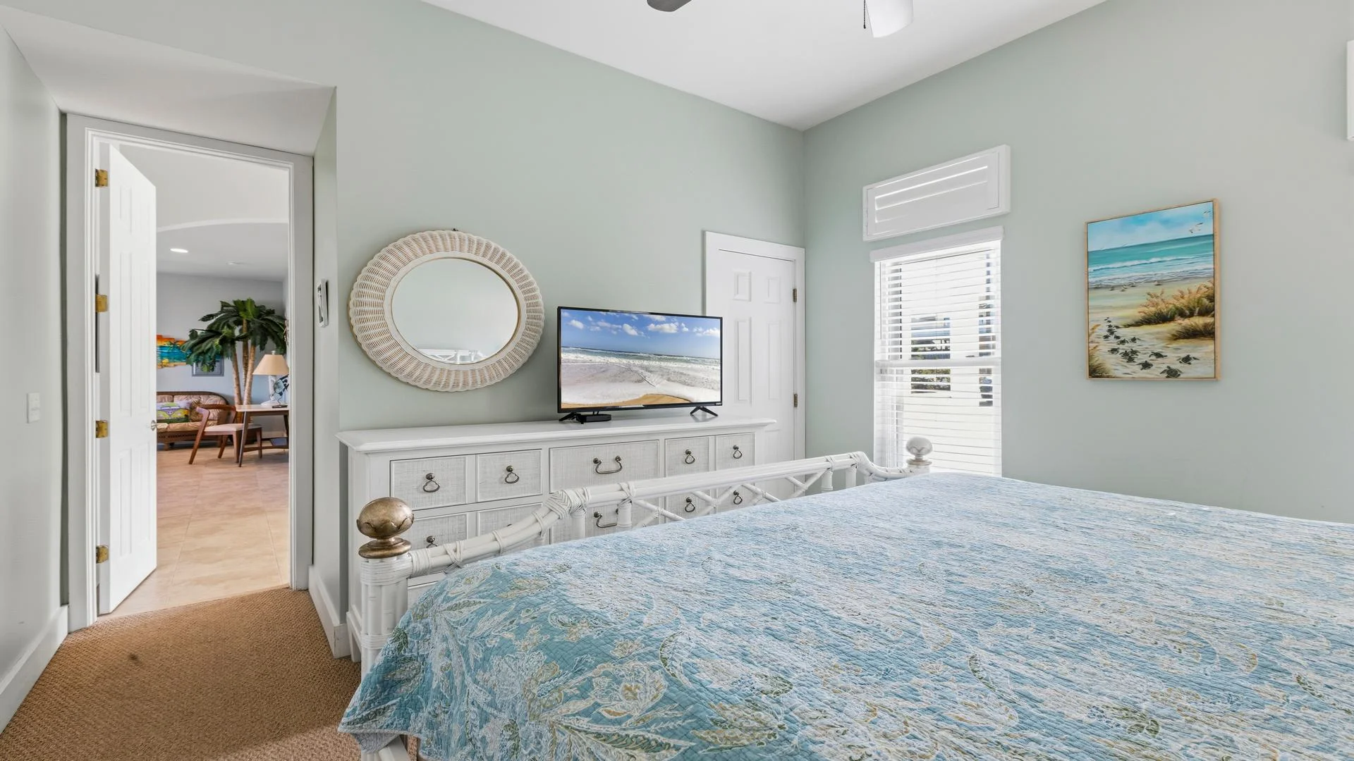 Bedroom with white rattan bed frame, blue floral quilt, dresser topped by round wicker mirror and flat-screen TV.
