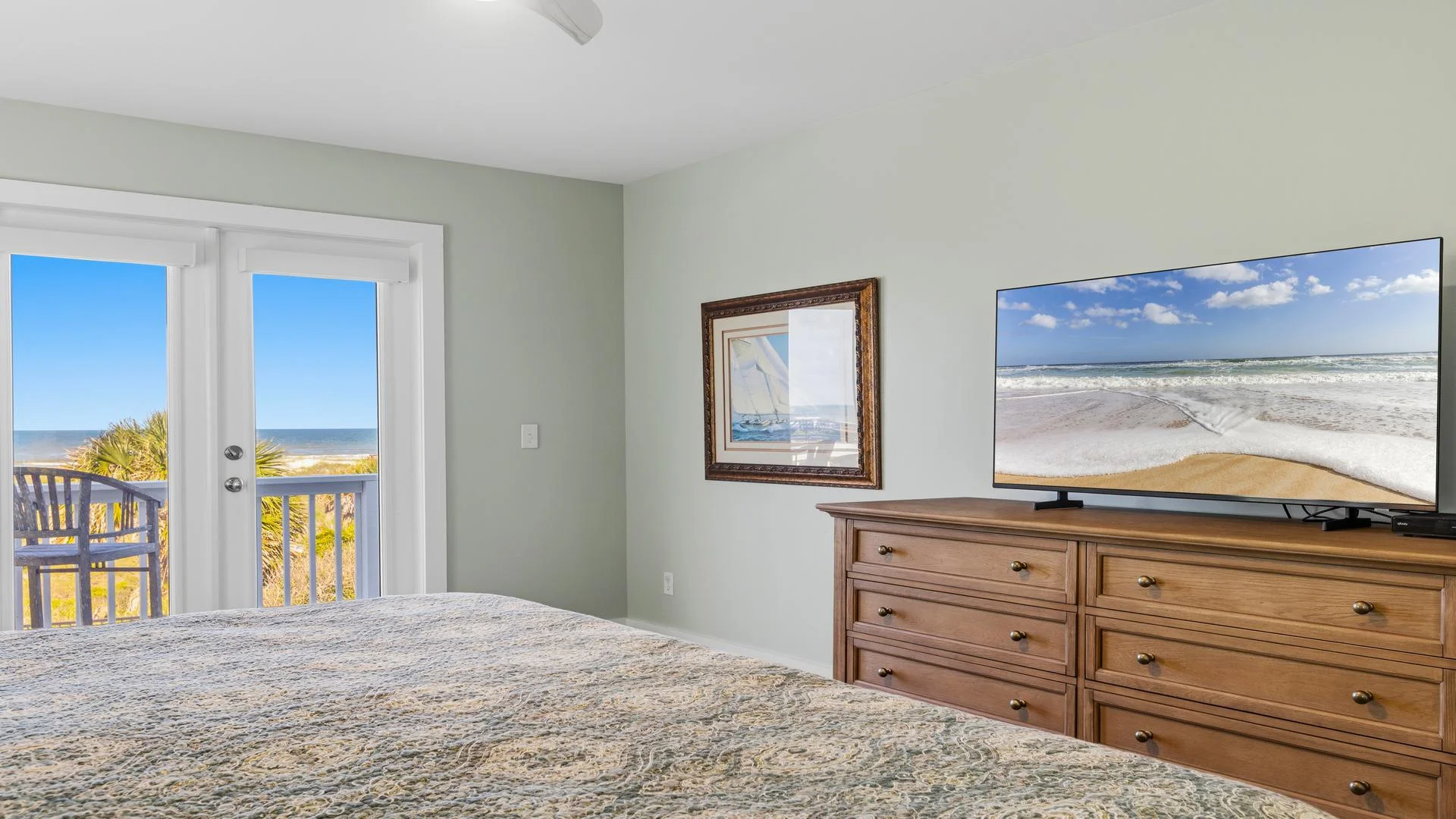 Bedroom features a wooden dresser with flat-screen TV and ocean views beyond glass doors leading to a balcony.