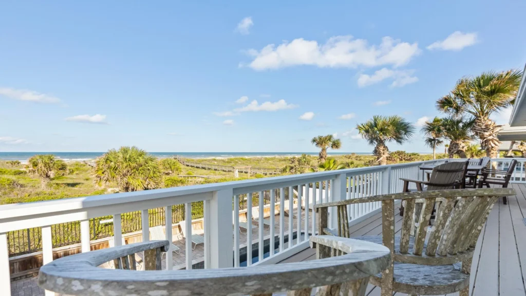 Oceanfront deck with weathered wooden Adirondack chairs, white railing, and palm trees framing the distant beach view.