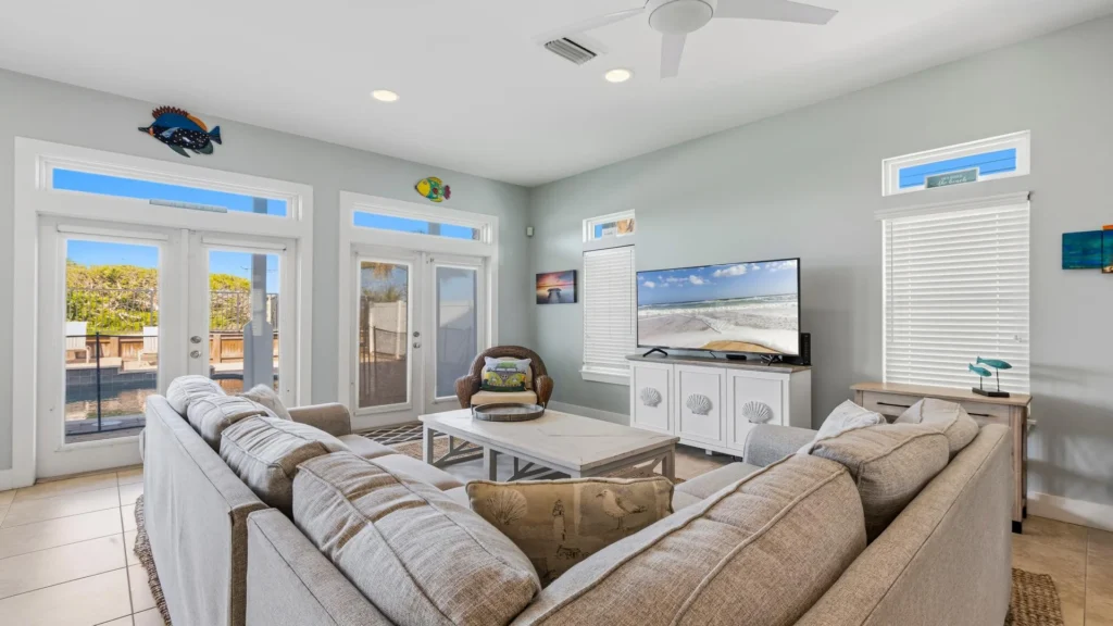 First-floor living room with a large gray sectional, wicker chair, white media console, and pool view through glass doors.