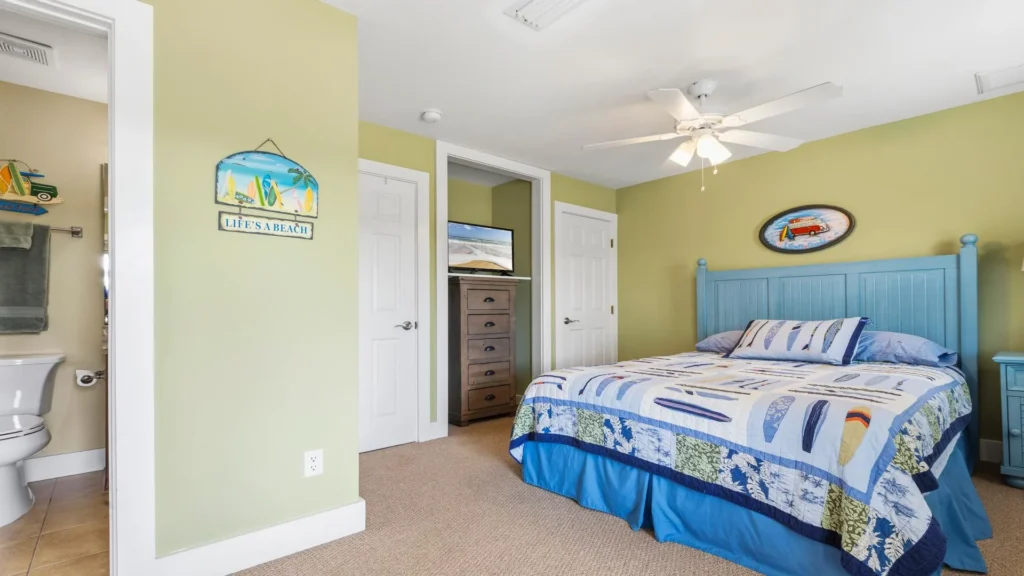 Second-floor bedroom with light green walls, blue wood headboard, surfboard-themed quilt, and TV on a wooden dresser.