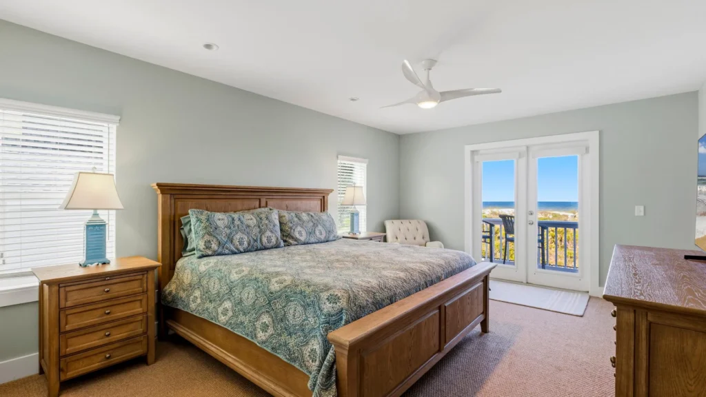 The second-floor primary bedroom features a wooden king bed with patterned blue bedding, matching nightstands, and ocean views.