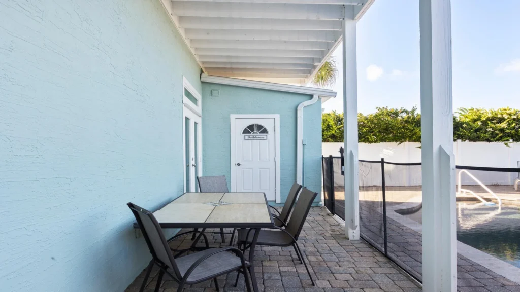Poolside patio with tiled stone floor, a square dining table with four gray mesh chairs, and a light blue bathhouse door.