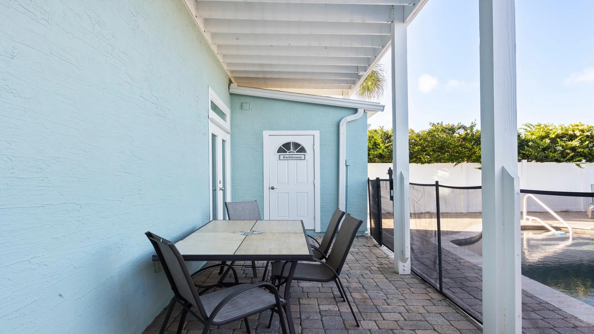 Poolside patio with tiled stone floor, a square dining table with four gray mesh chairs, and a light blue bathhouse door.