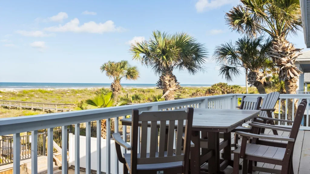Oceanfront balcony deck with wooden dining table and chairs, overlooking palm trees and sandy dunes.