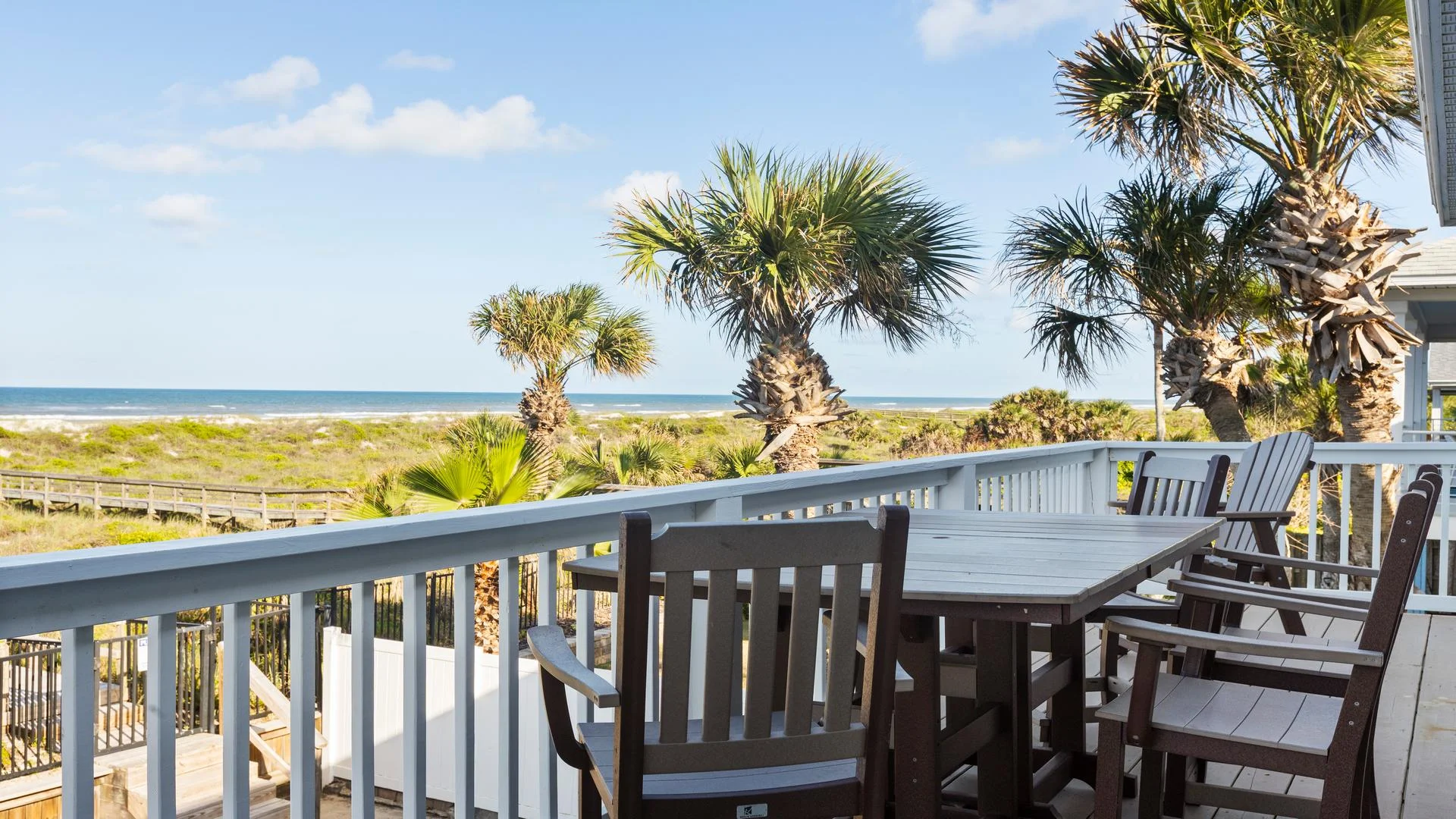 Oceanfront balcony deck with wooden dining table and chairs, overlooking palm trees and sandy dunes.