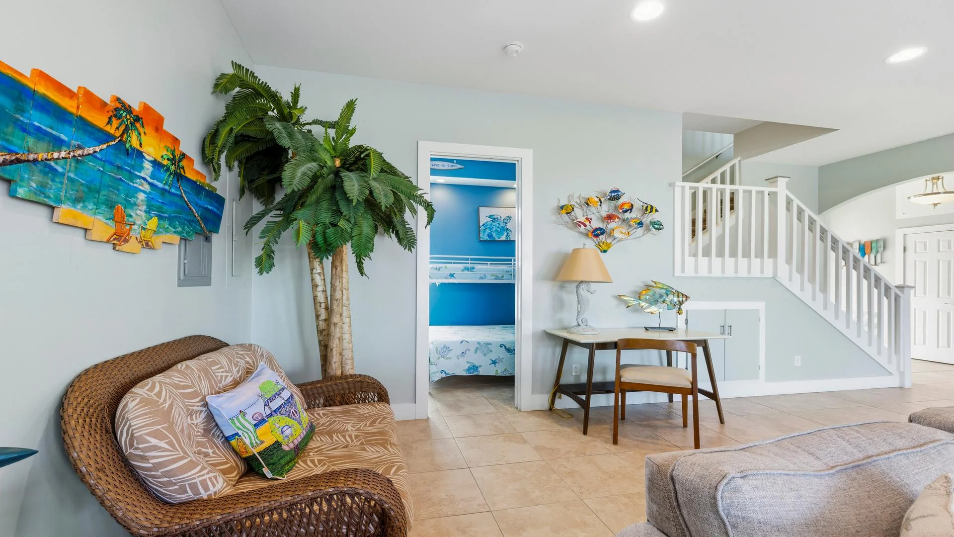 Wicker loveseat with leaf-print cushions sits beside a tropical wall art and faux palm tree in the living area.