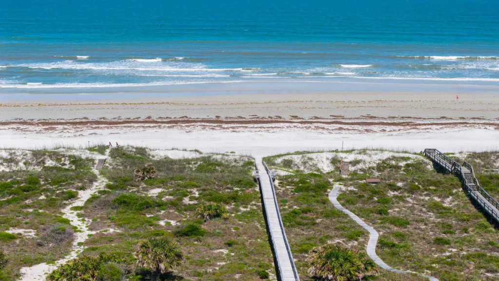 Wooden boardwalk and sandy paths lead through coastal dunes to a wide beach with gentle ocean waves.