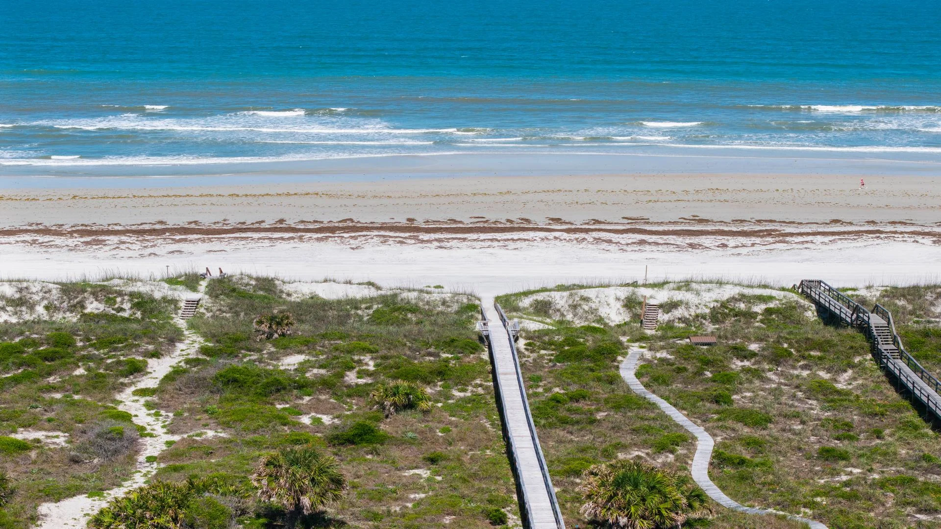 Wooden boardwalk and sandy paths lead through coastal dunes to a wide beach with gentle ocean waves.