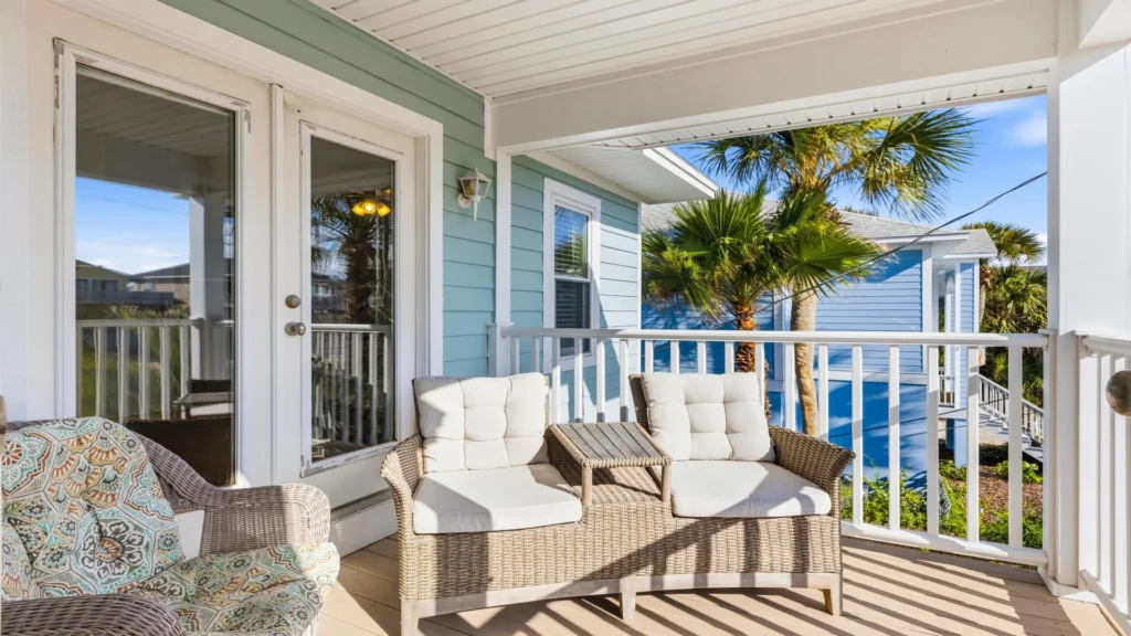 Covered porch with cushioned wicker loveseat and armchair, overlooking palm trees and neighboring blue house.