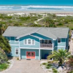 Oceanfront exterior of a blue two-story house with coral double doors, balcony seating, and sandy beach access.