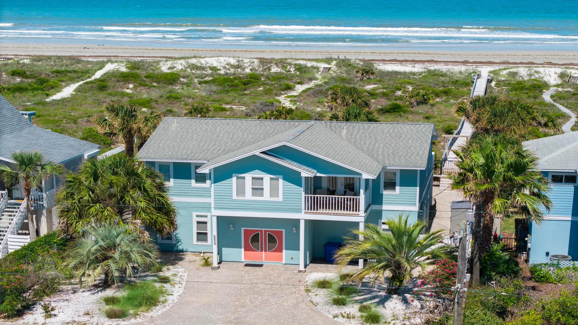 Oceanfront exterior of a blue two-story house with coral double doors, balcony seating, and sandy beach access.
