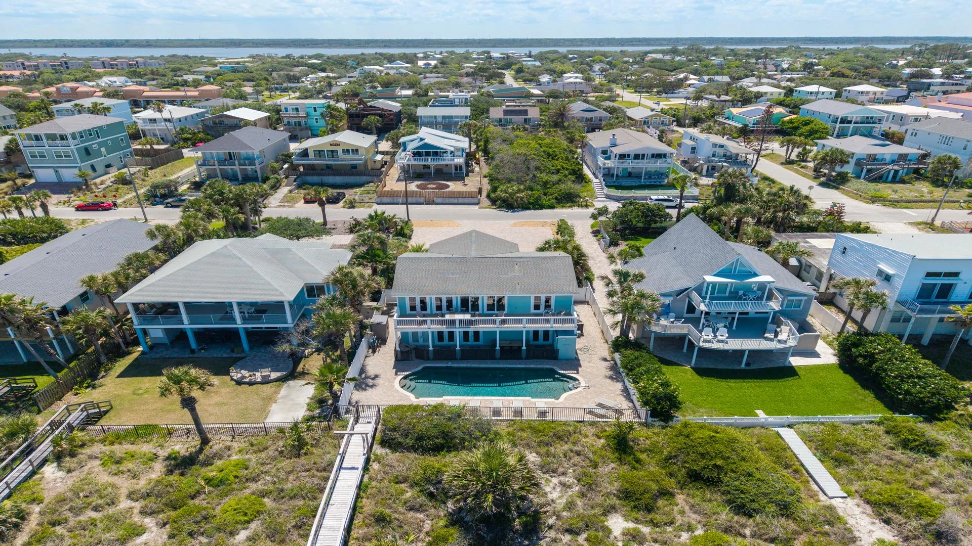 Aerial view of oceanfront house with a pool, upper and lower balconies, sandy beach access, and lush dune vegetation.