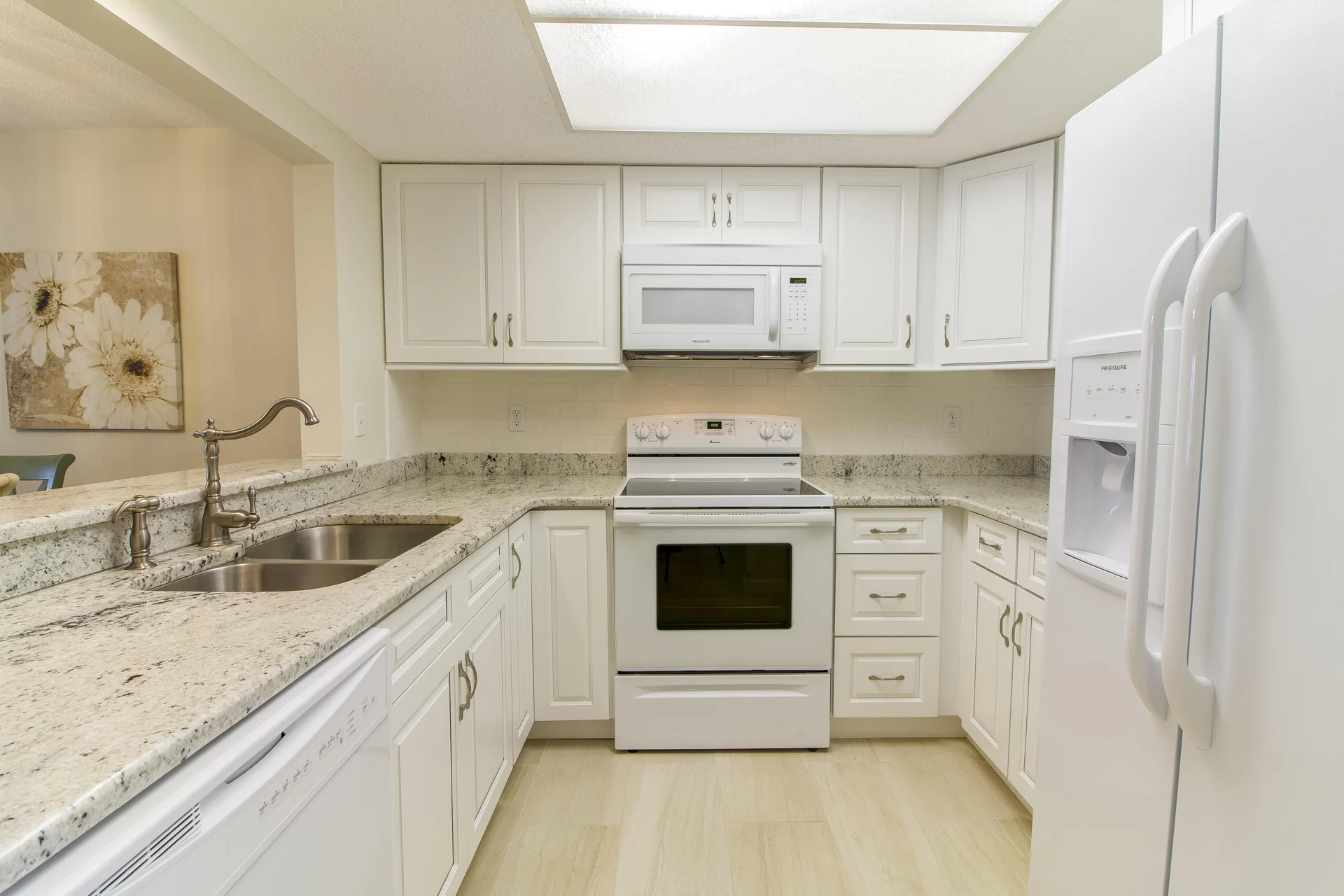 U-shaped kitchen with white cabinetry, granite countertops, double sink, and white appliances under soft ceiling light.