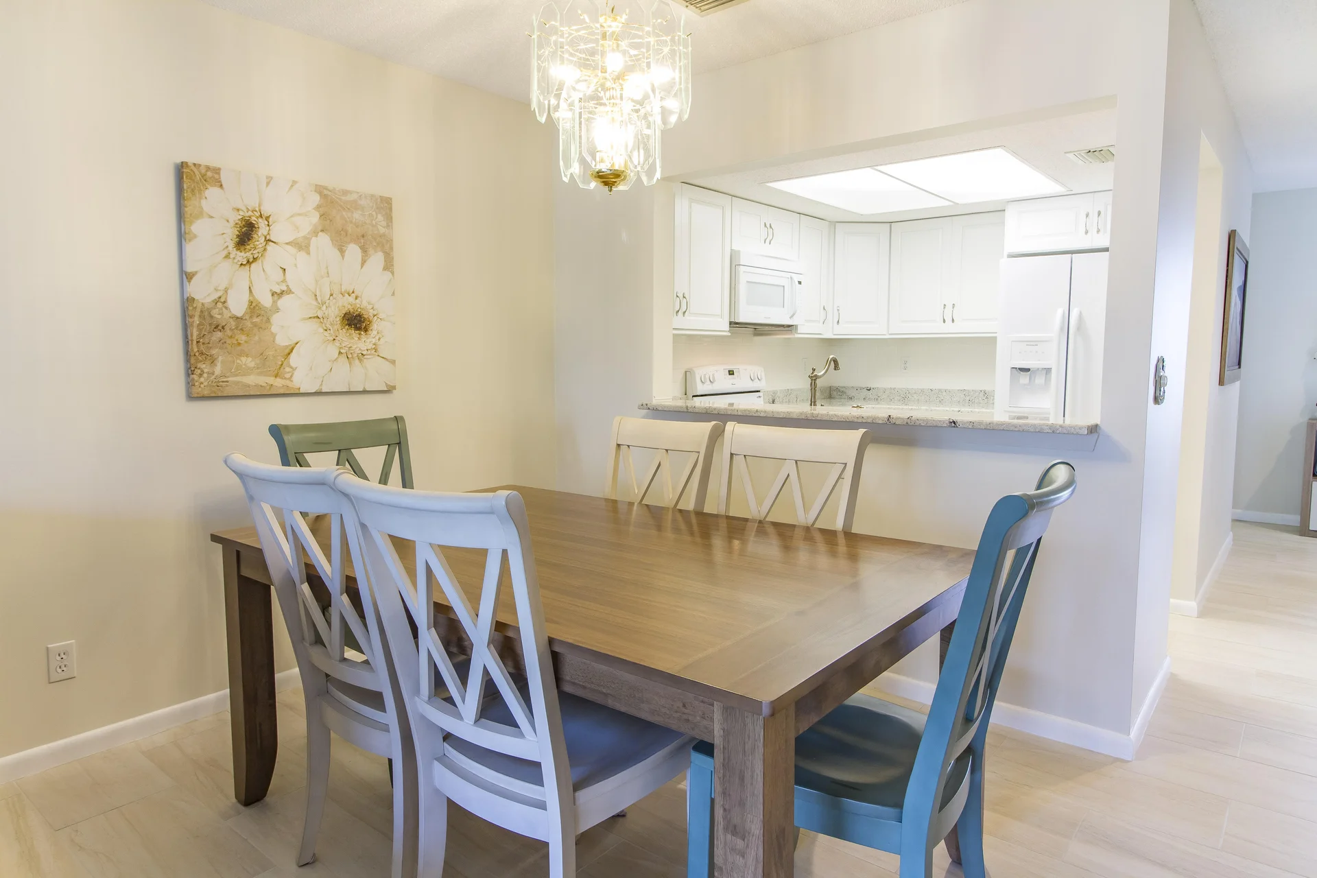 Dining area with wooden table, six mismatched chairs, floral wall art, and a glass chandelier above.