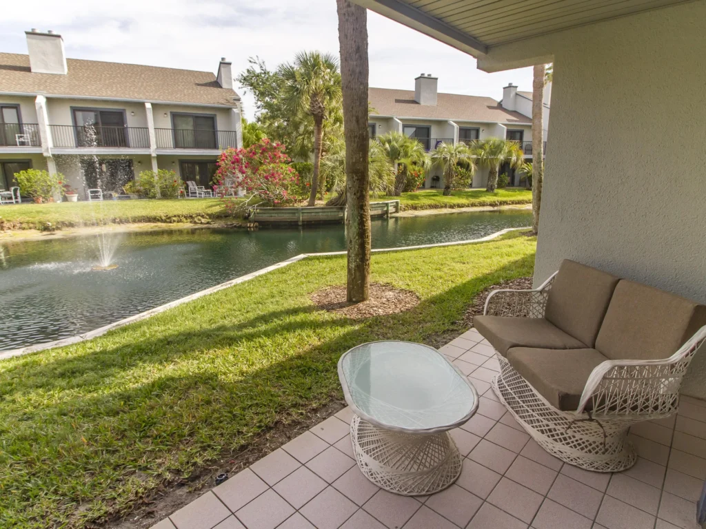 Covered patio with white wicker loveseat and glass-top table overlooking lagoon with fountain and palm trees.