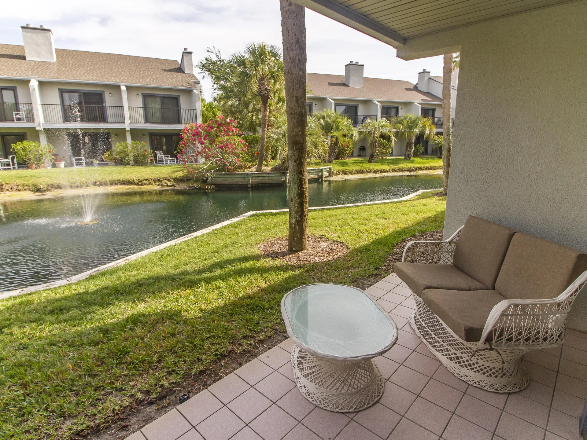 Covered patio with white wicker loveseat and glass-top table overlooking lagoon with fountain and palm trees.