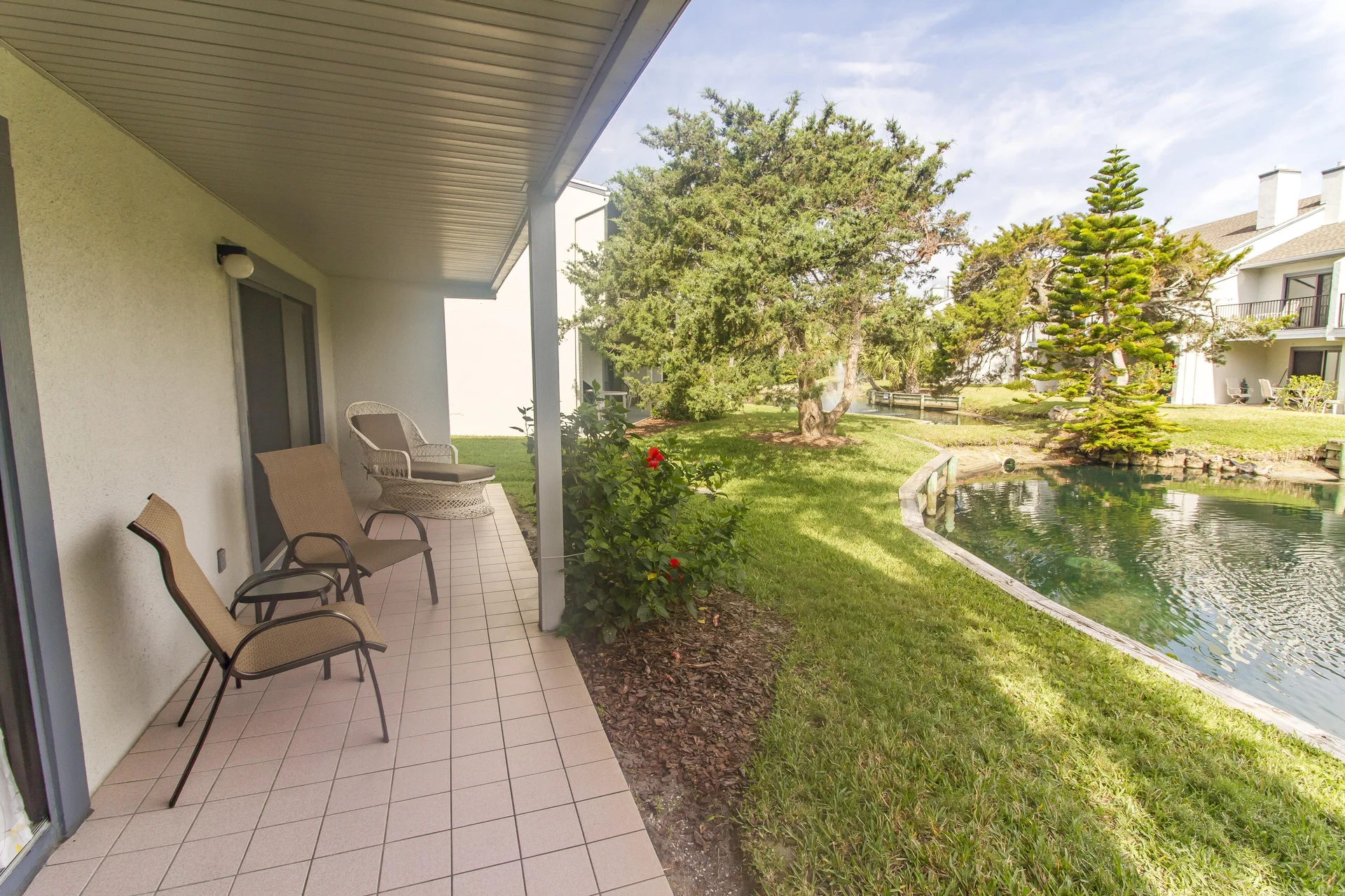 Ground floor patio with beige tile flooring, metal-framed chairs, wicker lounge, and lagoon water view.
