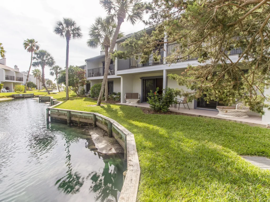Patio features white wicker seating and small table nestled beneath shaded trees beside a tranquil lagoon.