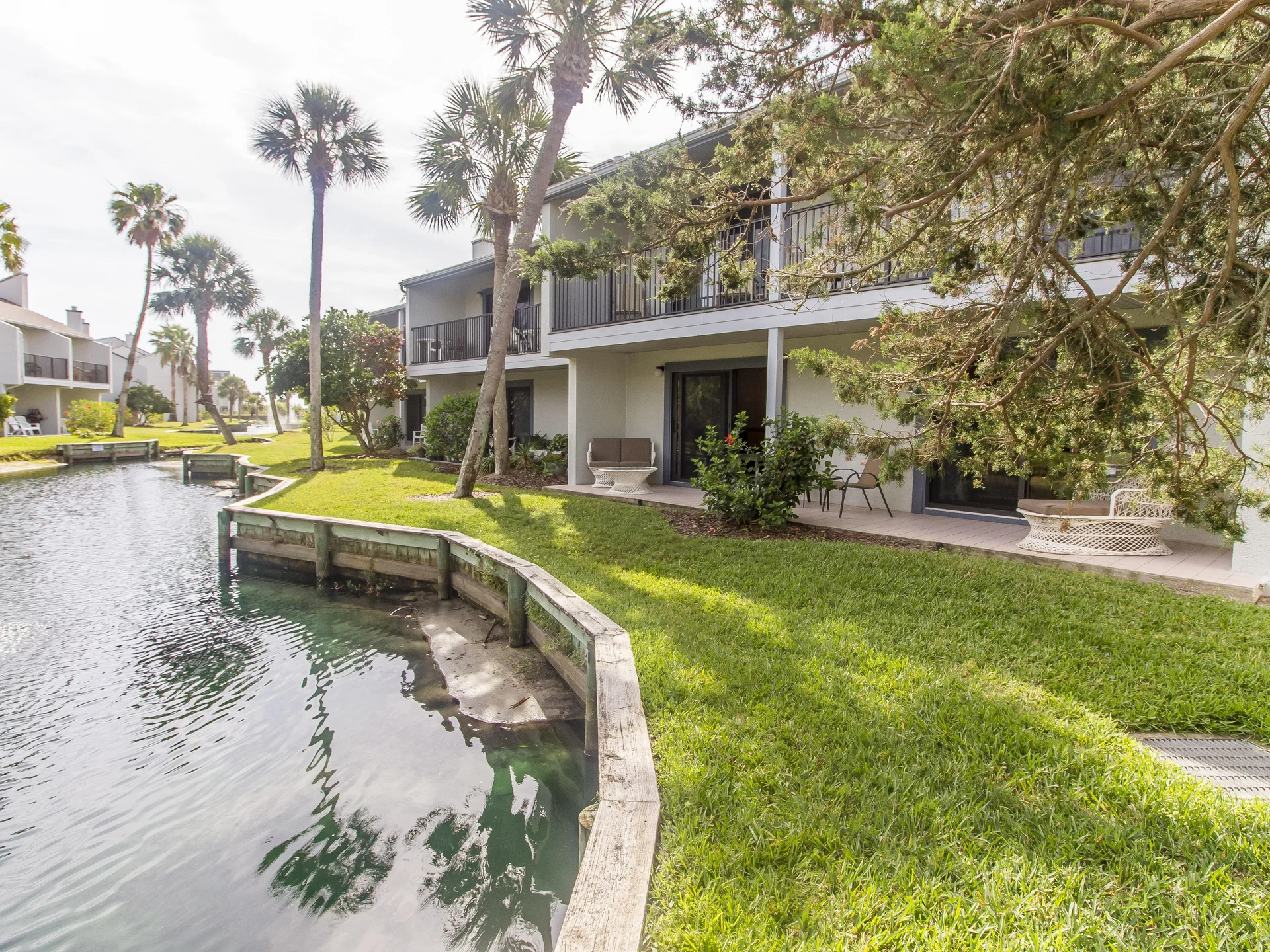 Patio features white wicker seating and small table nestled beneath shaded trees beside a tranquil lagoon.