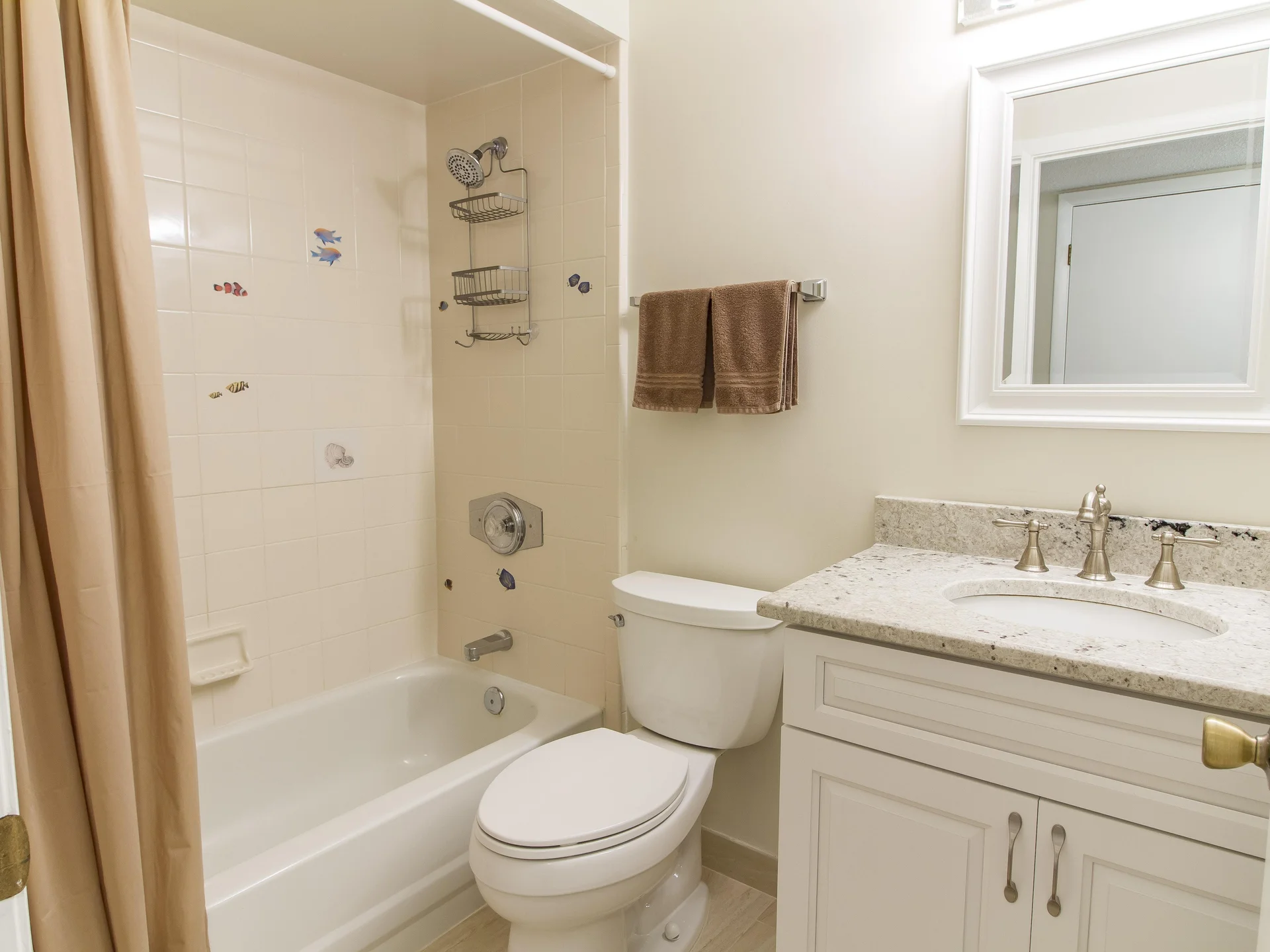 Tub and shower combo with beige curtain, fish-themed tile accents, white toilet, and granite-topped vanity with framed mirror.