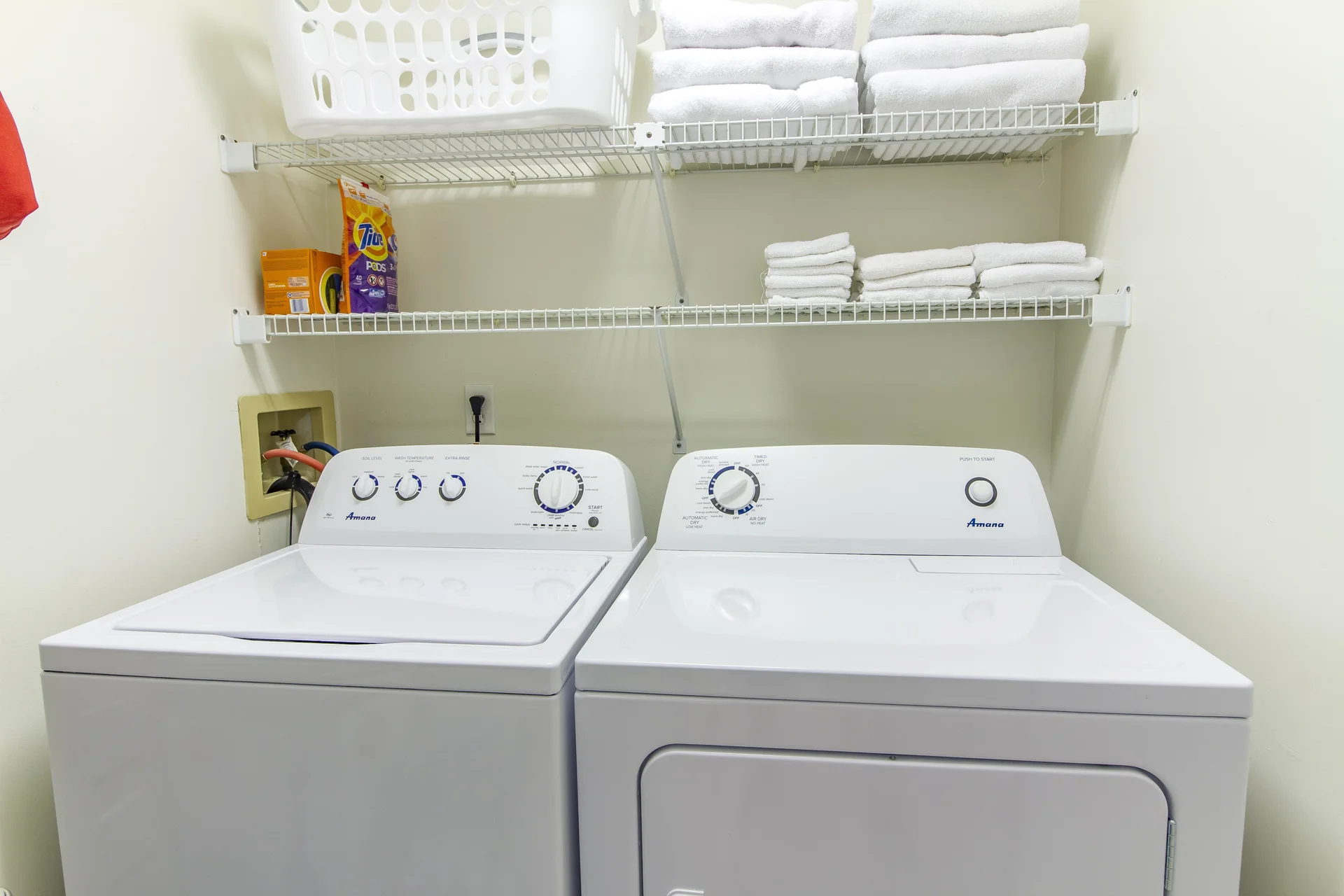 Laundry area with white top-load washer and dryer beneath wire shelving stocked with folded white towels.
