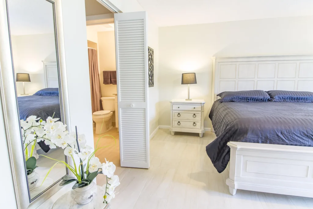 Bedroom with white wood bedframe, matching nightstand, black lamp, and full-length mirror reflecting orchid arrangement.