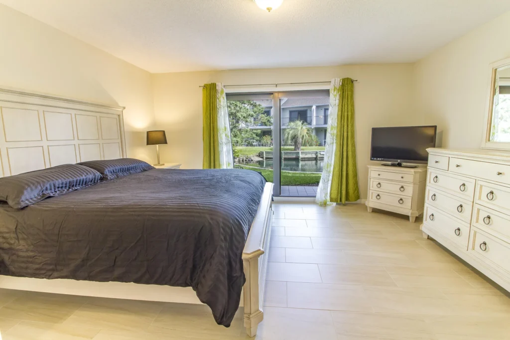 Master bedroom with white wooden bedframe, black bedding, matching dresser and TV, sliding doors to lagoon view.