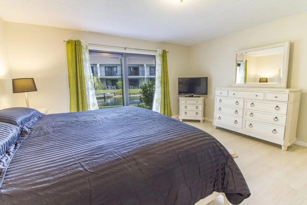 Master bedroom features a dark striped bedspread, white dresser with mirror, and sliding glass doors with lagoon views.