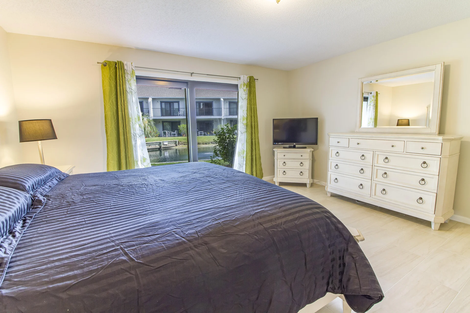 Master bedroom features a dark striped bedspread, white dresser with mirror, and sliding glass doors with lagoon views.