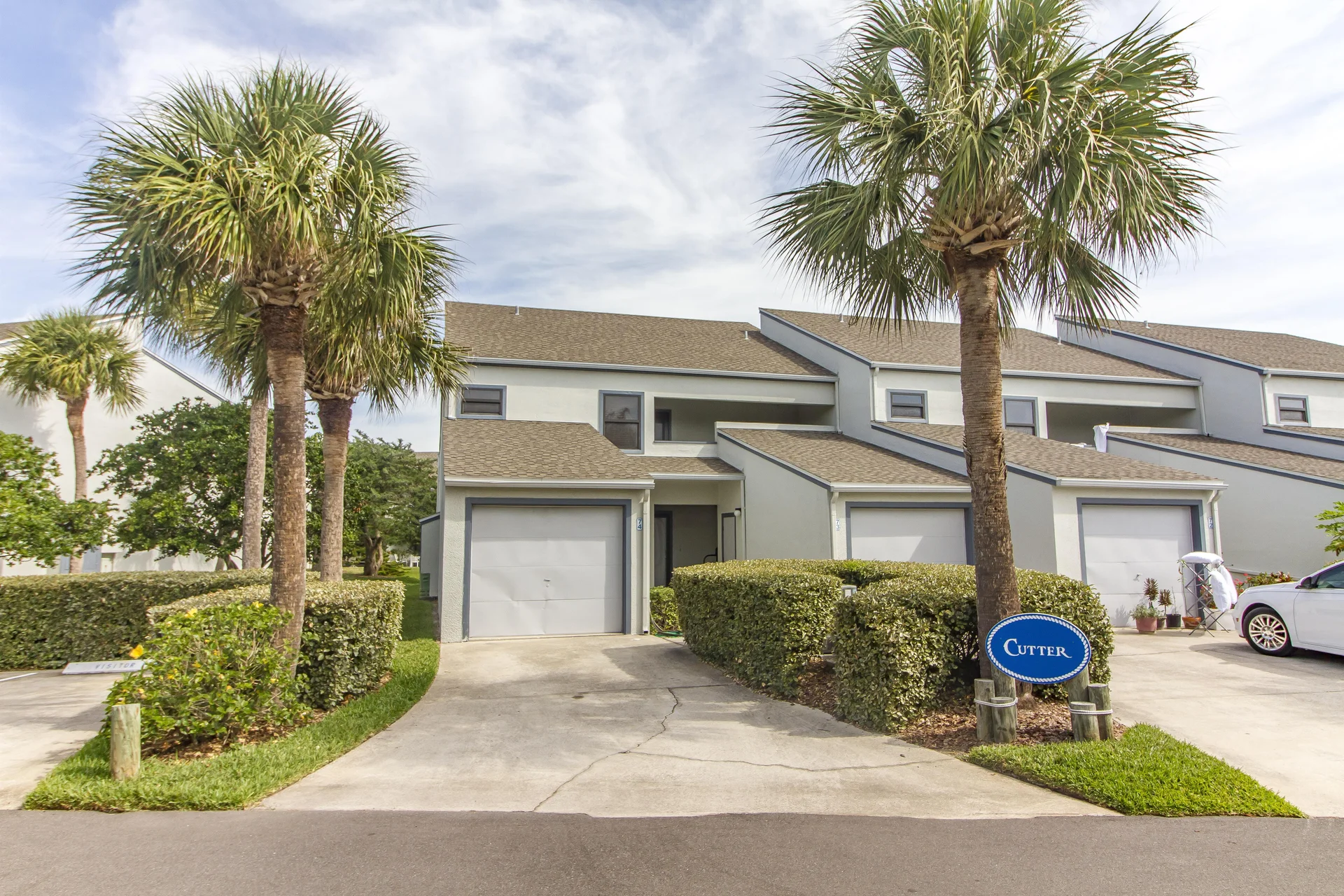 Exterior view of a two-story condo with attached garage, beige siding, and a driveway framed by palm trees and hedges.