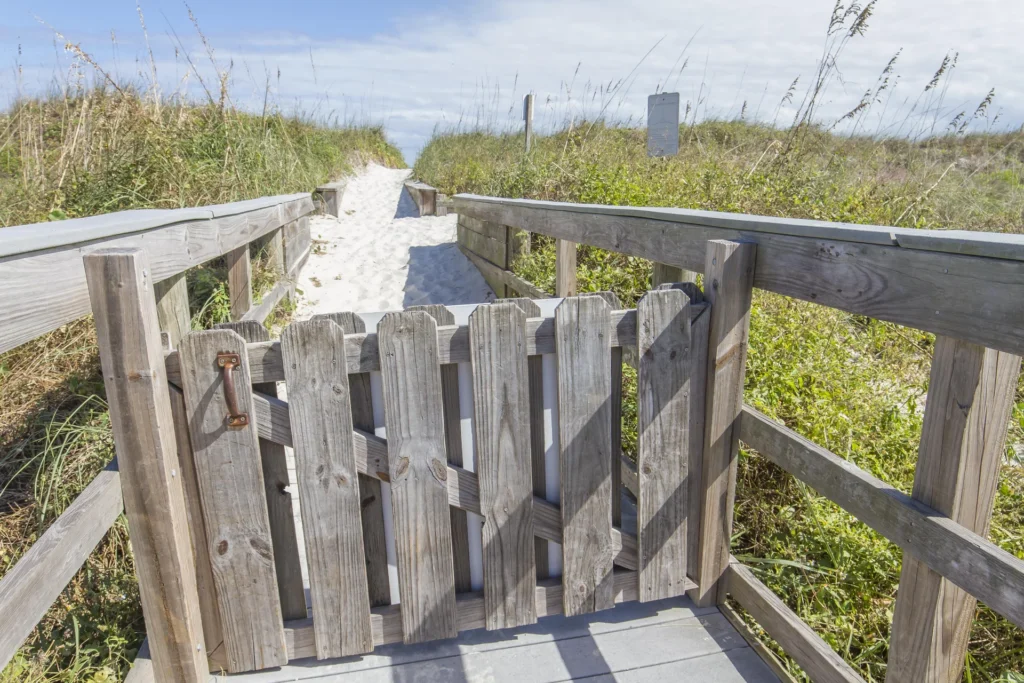 Wooden gate opens to a sandy path flanked by grassy dunes under a blue sky.