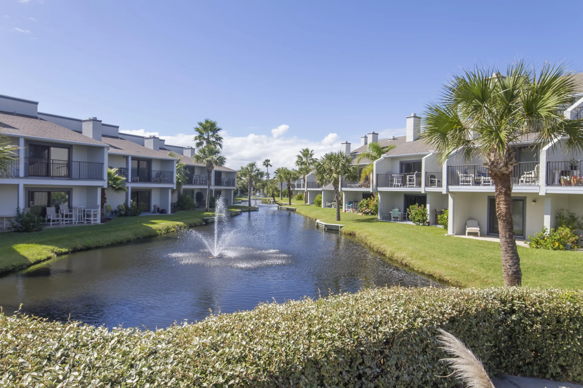 Lagoon view between two rows of two-story condos with patios, palm trees, and a central water fountain.