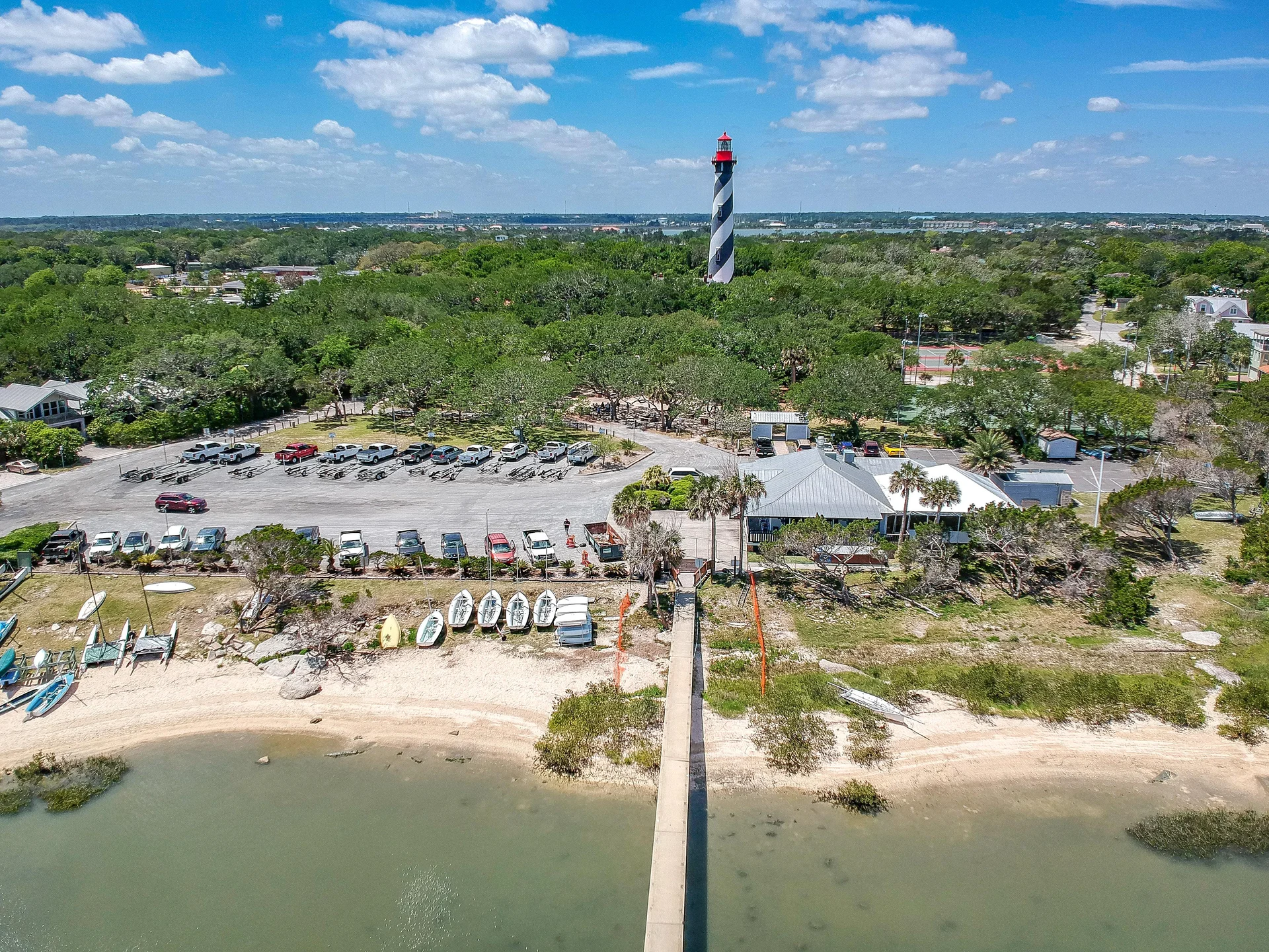 Aerial view of sandy shoreline with dock, parked boats, adjacent parking lot, and tall black-and-white lighthouse beyond.