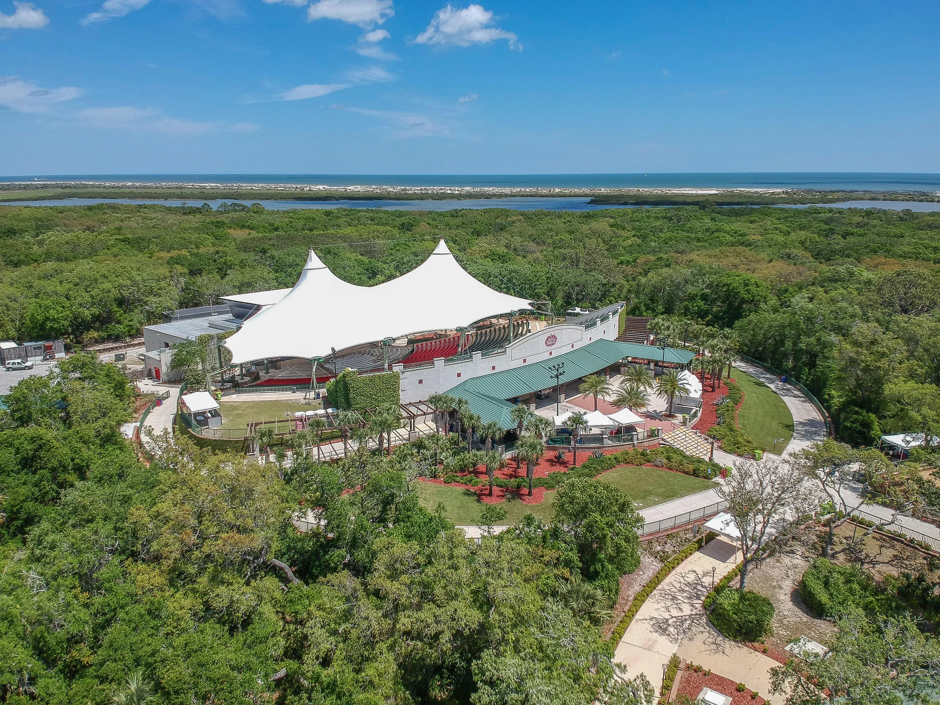 Aerial view of a white tensile roof covering outdoor seating, surrounded by lush greenery and a distant ocean horizon.