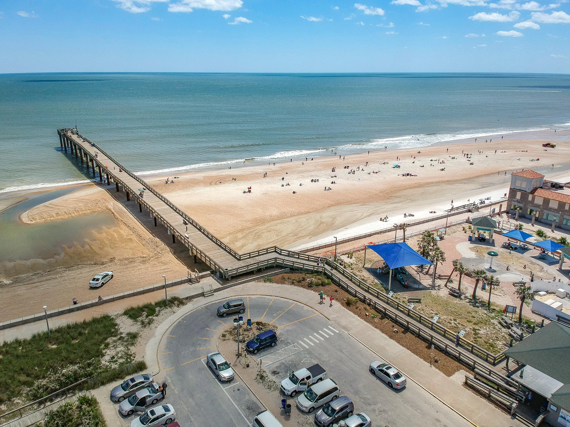 Aerial view of sandy beach with pier stretching into calm ocean, adjacent parking lot, and shaded pavilion area.