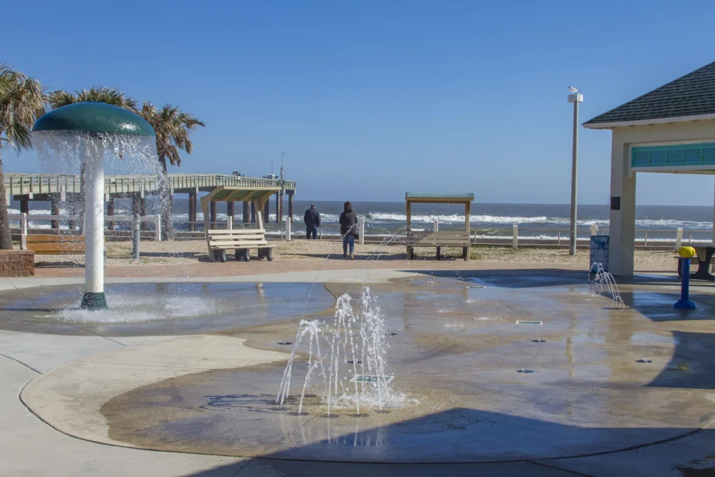 Outdoor splash pad with water jets and mushroom fountain, sandy beach, wooden benches, and ocean pier beyond.