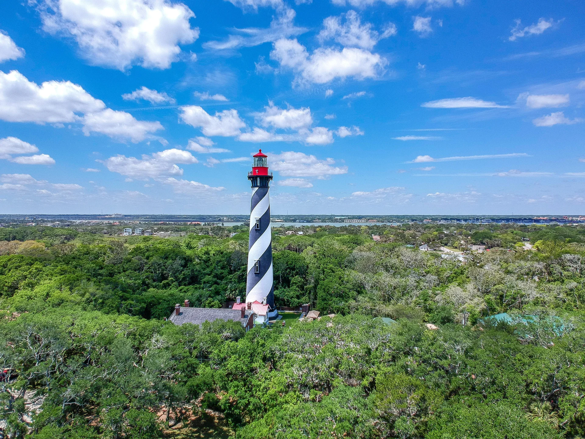 Aerial view of the black-and-white spiral lighthouse surrounded by dense green trees under a bright blue sky.