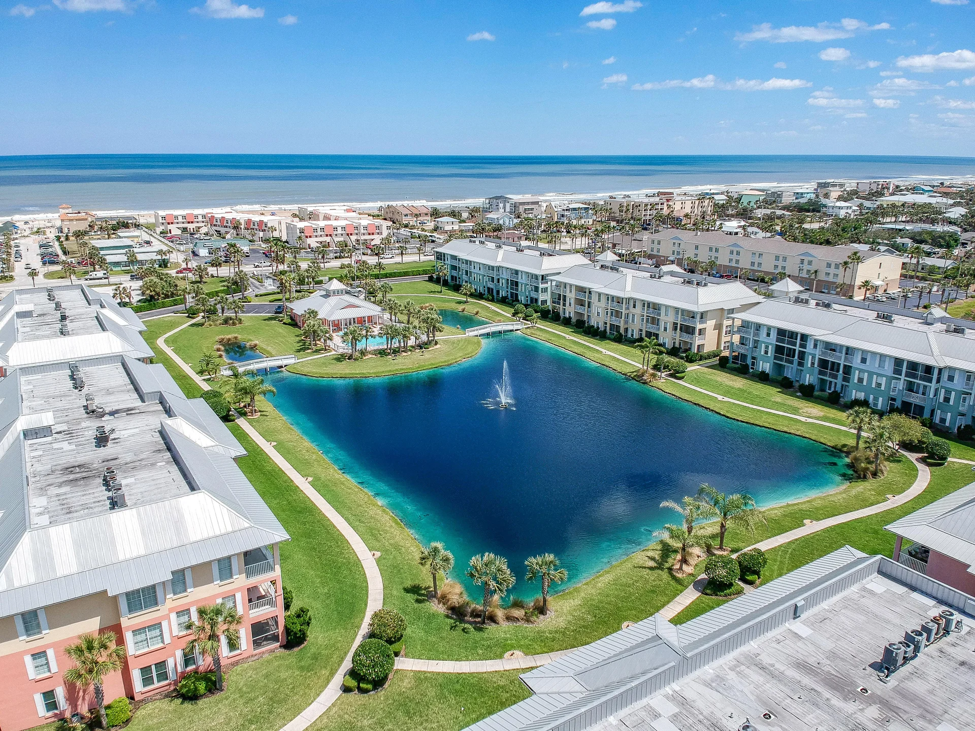 Aerial view of condo buildings surrounding a large blue pond with a central fountain and walking paths.