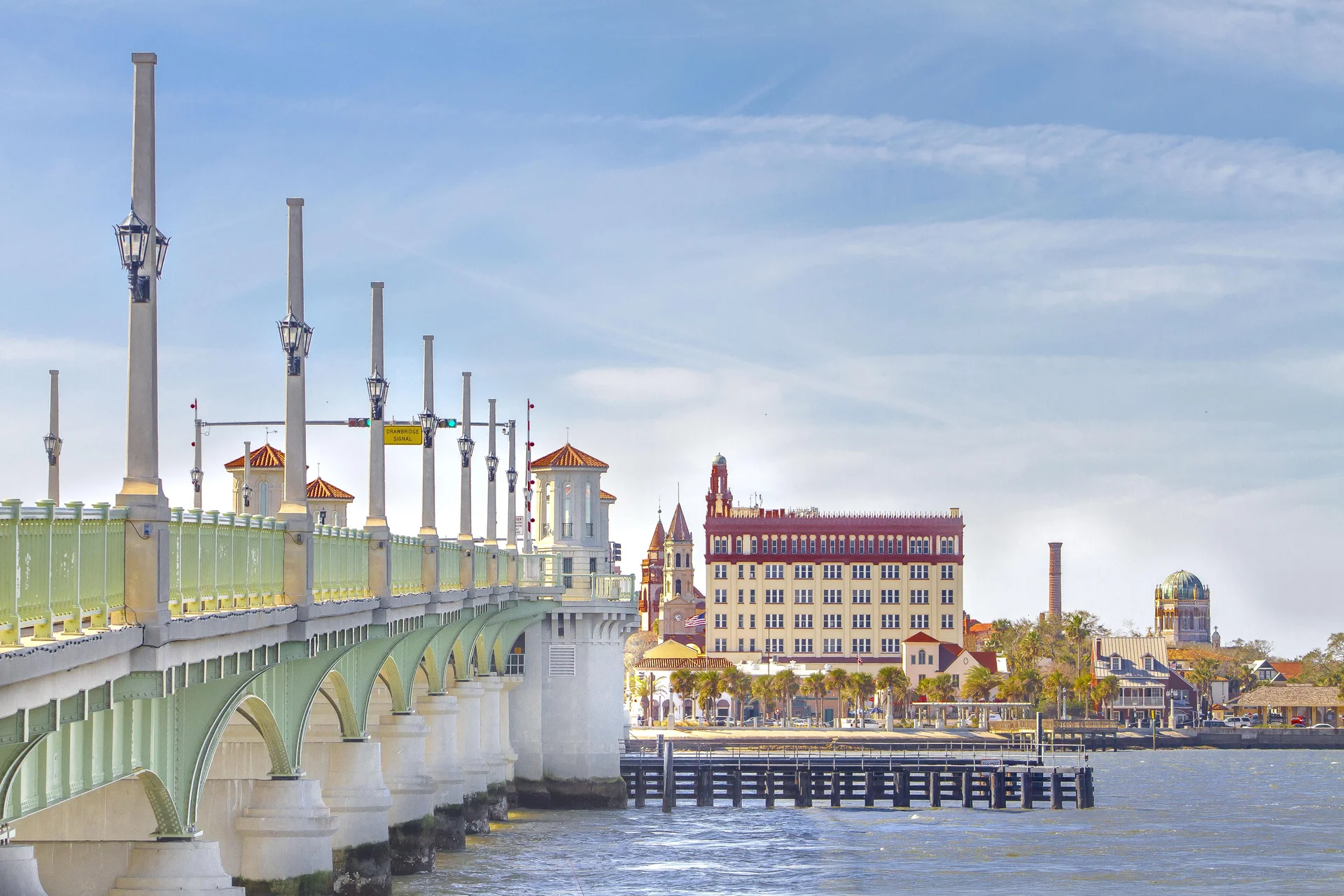 Historic bridge with green railings and tall lampposts stretches across blue water toward ornate buildings.