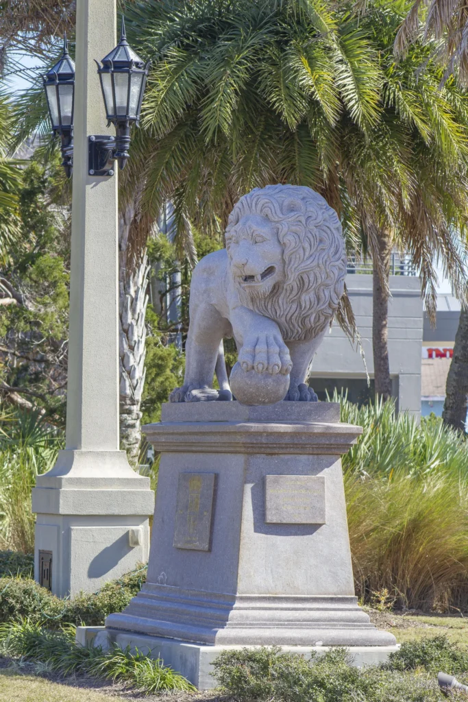 Outdoor stone lion statue with detailed mane and paw resting on a sphere, surrounded by palm trees and greenery.