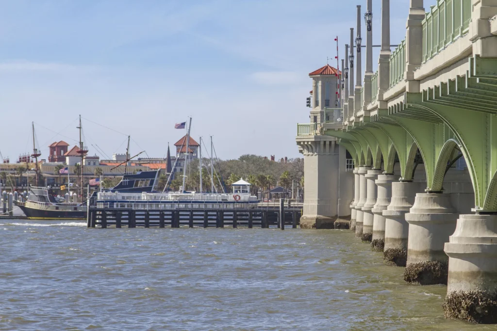 View from the water showing the green-arched bridge with concrete pillars and historic ships docked near a red-roofed waterfront.