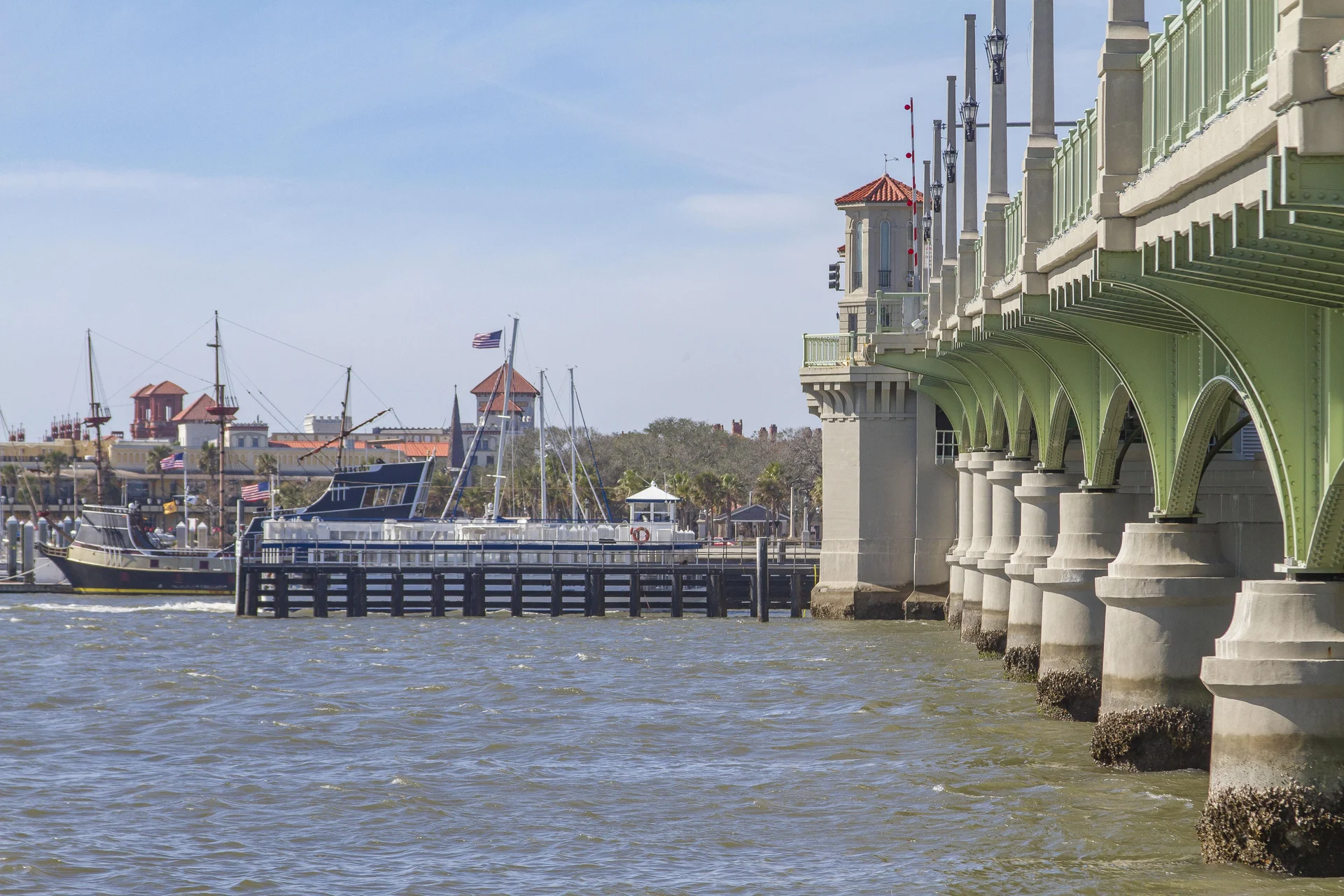 View from the water showing the green-arched bridge with concrete pillars and historic ships docked near a red-roofed waterfront.