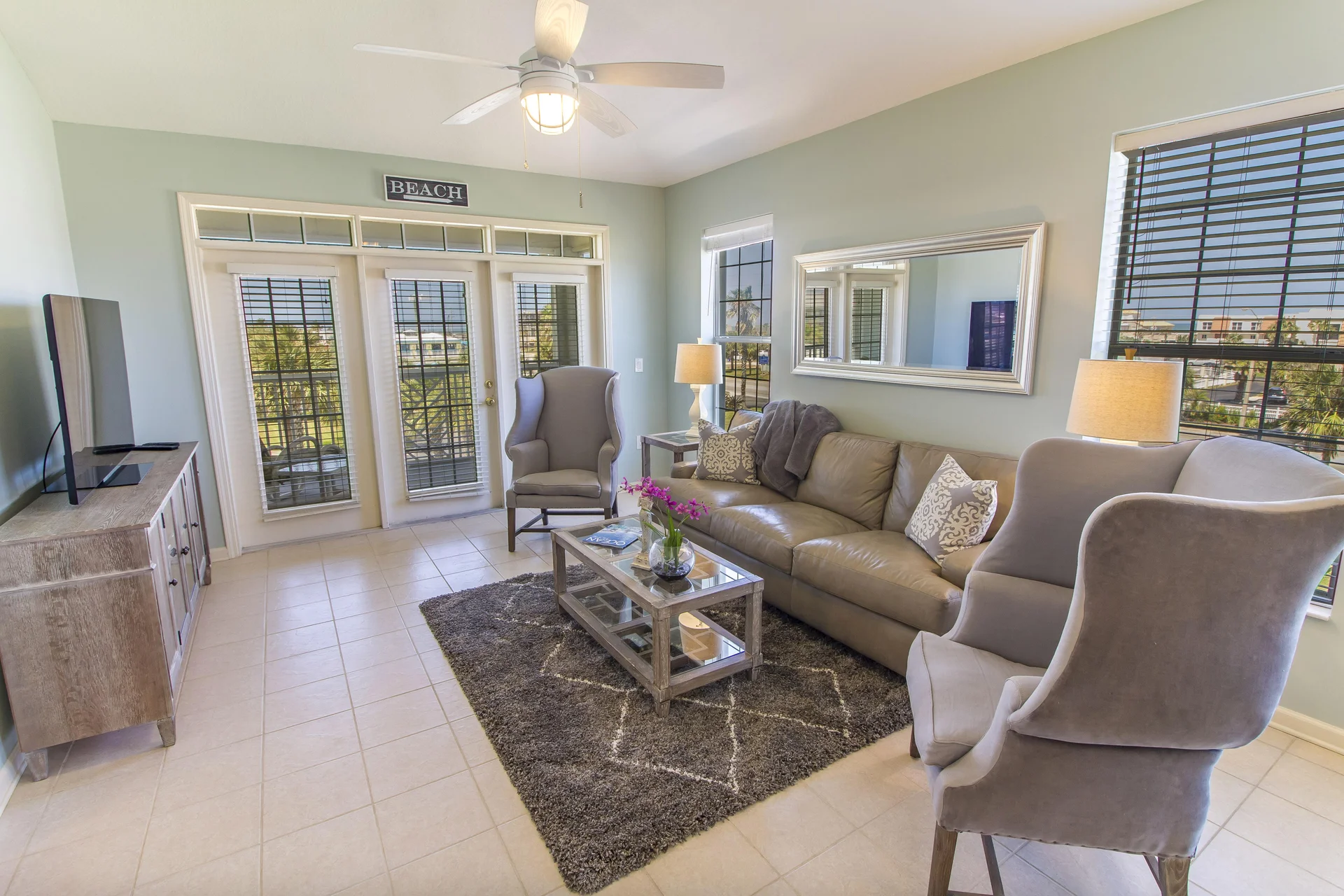 Soft sage walls frame the living room with plush gray wingback chairs, a taupe leather sofa, and tiled floors.