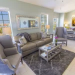 Living room with gray wingback chairs, leather sofa, glass-top coffee table on patterned rug, and dining area beyond.
