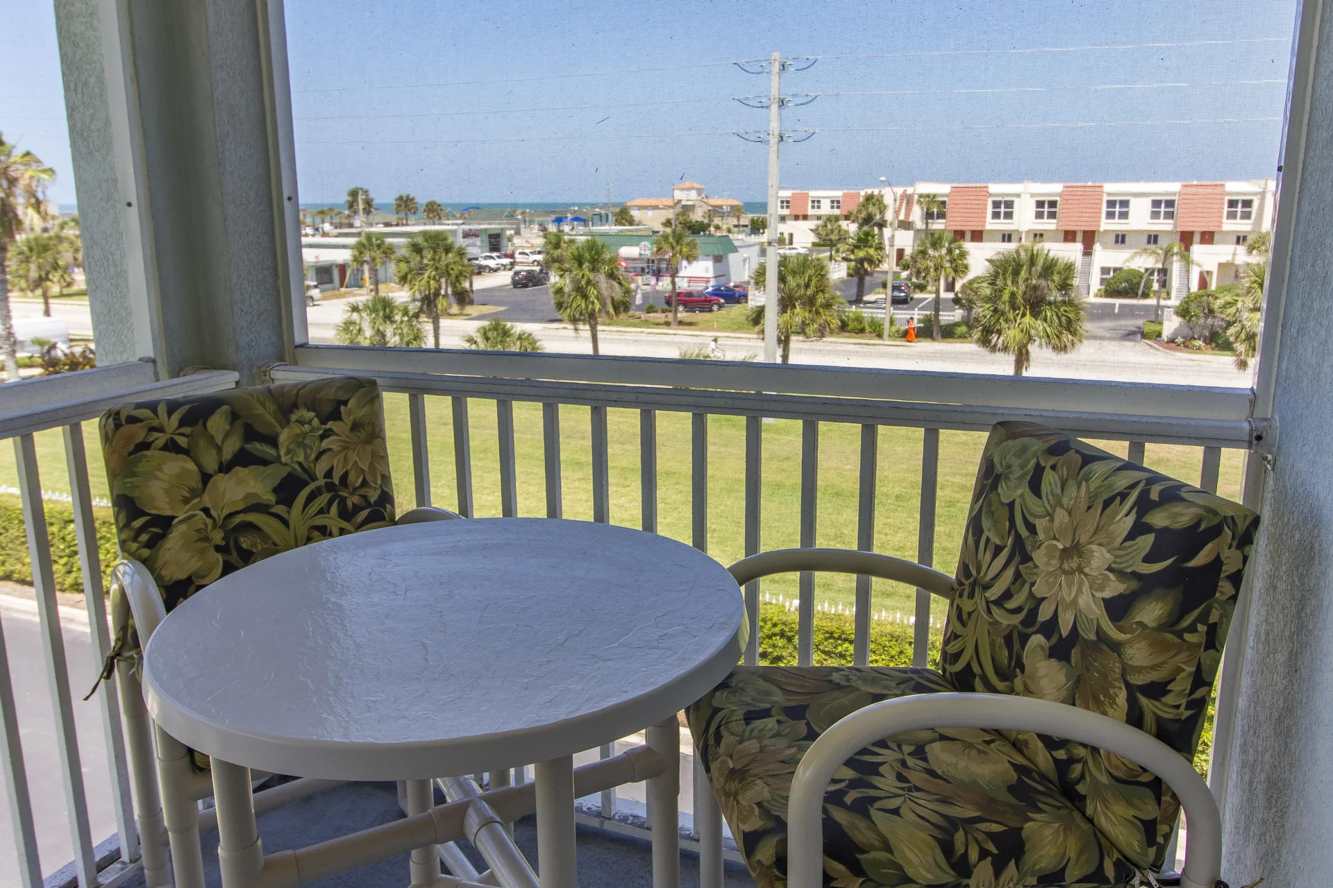 Screened balcony with white round table and cushioned chairs overlooking palm trees and distant ocean.