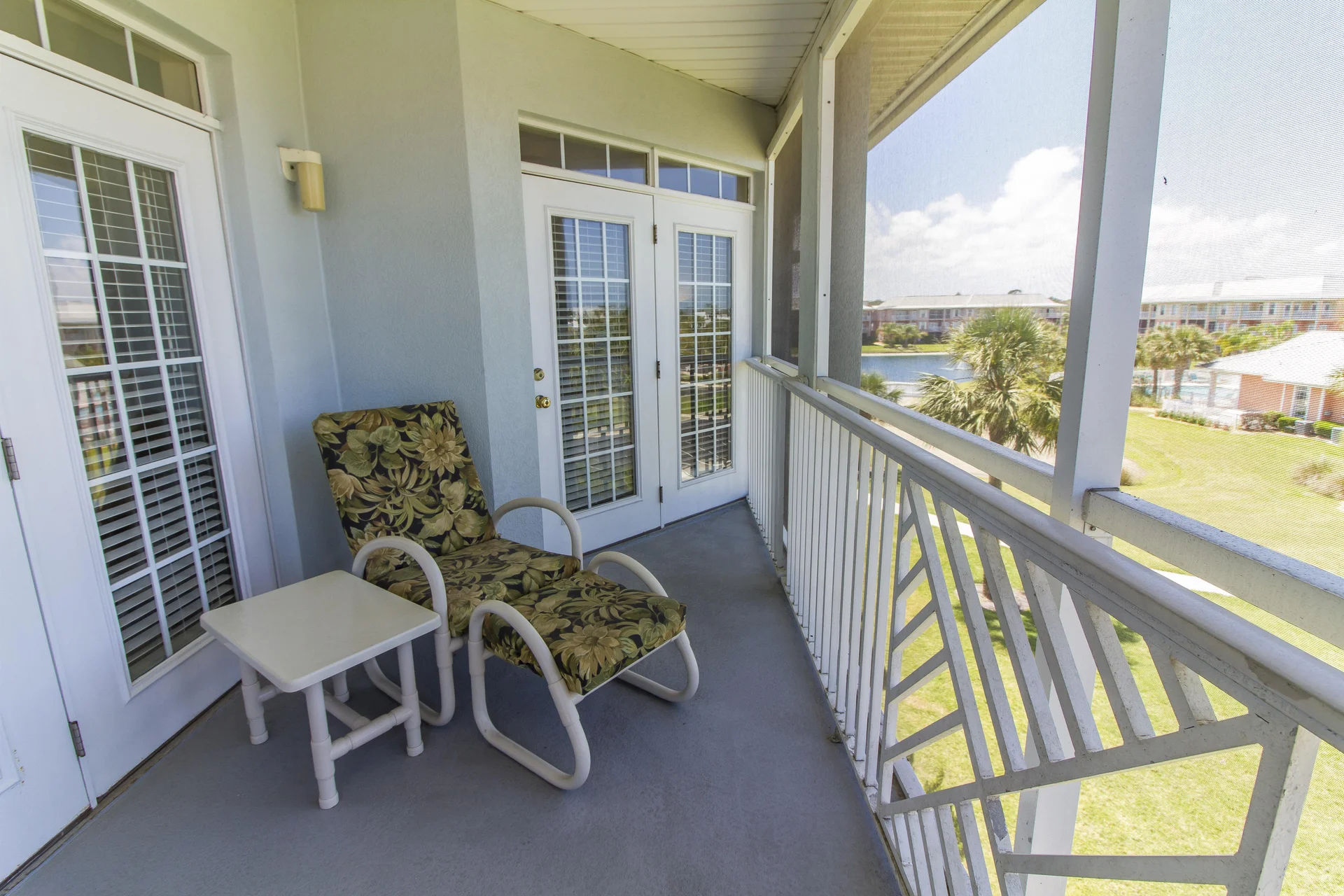 Outdoor balcony with cushioned floral lounge chair, white side table, screened railing, and views of landscaped grounds.
