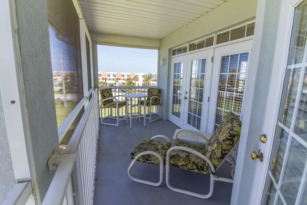 Outdoor balcony with floral cushioned lounge chairs, small round dining table, white railings, and screened walls.
