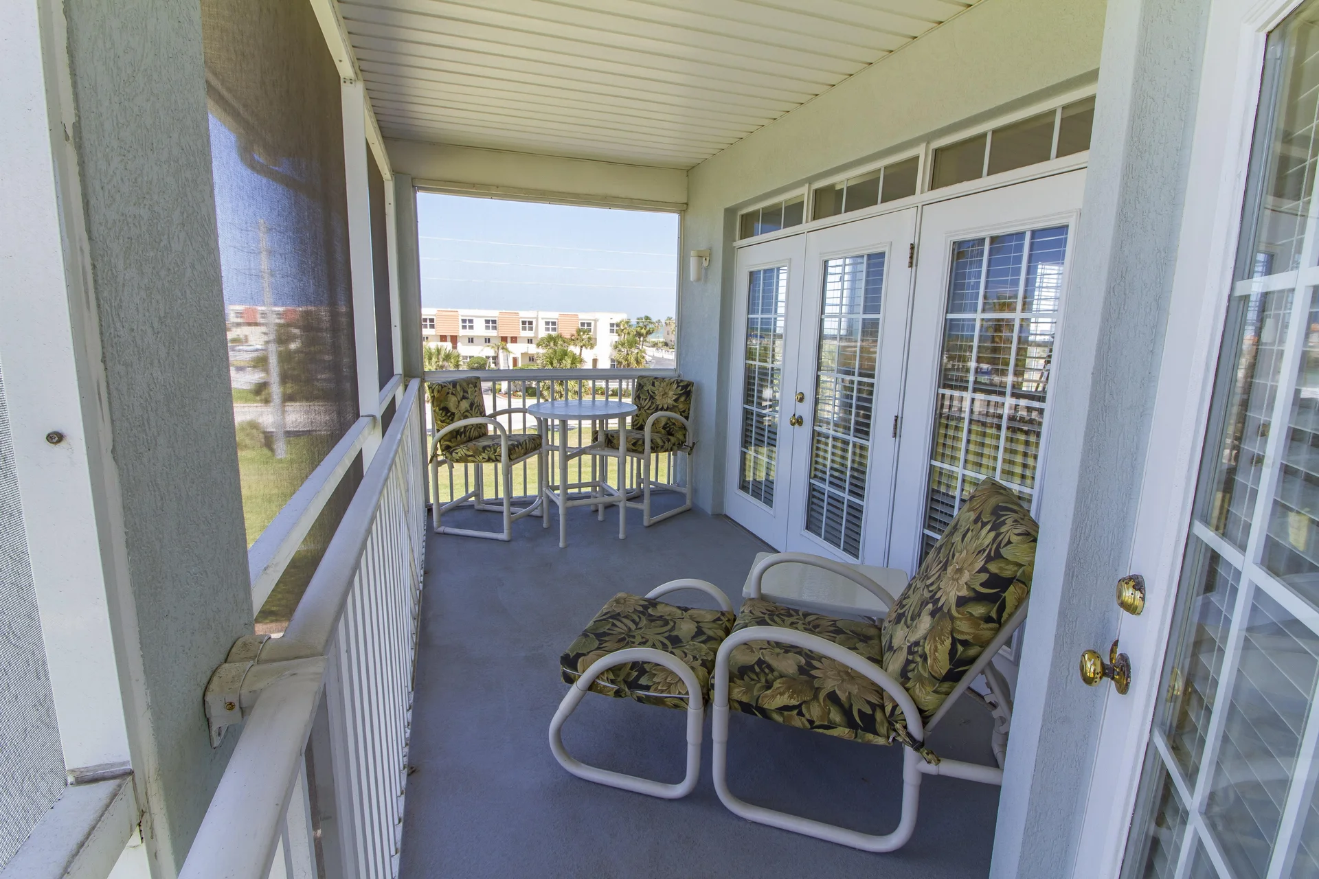 Outdoor balcony with floral cushioned lounge chairs, small round dining table, white railings, and screened walls.