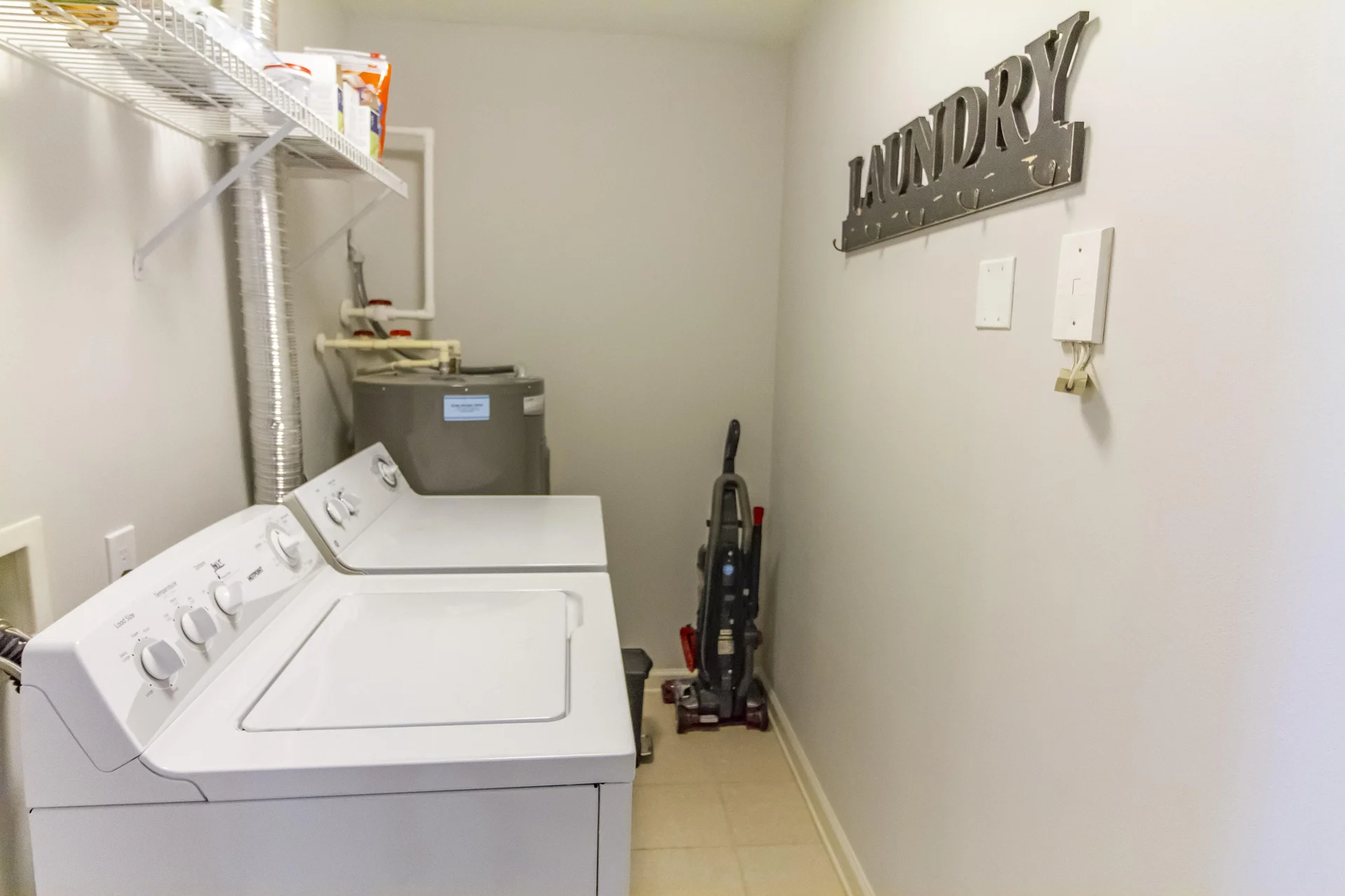Laundry room with white washer and dryer, beige tiled floor, vacuum cleaner, and metal "Laundry" wall sign.